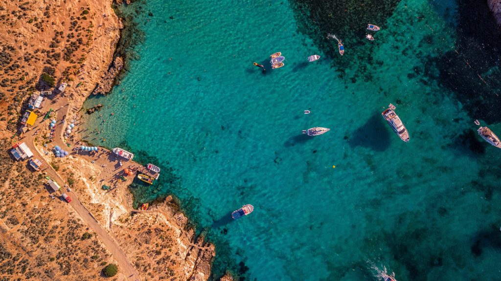 Aerial view of a rocky coastline with turquoise water, boats in the bay, and a small dock lined with vacation rentals and vehicles. The clear sea reveals dark patches of underwater rocks near the managed properties along the shore.