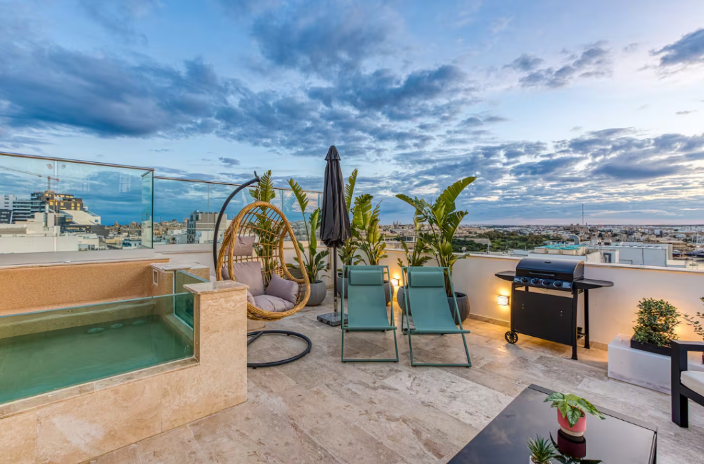 A rooftop terrace at sunset, ideal for vacation rentals, features a small pool, two lounge chairs, a hanging chair, potted plants, a barbecue grill, and cityscape views under a partly cloudy sky.