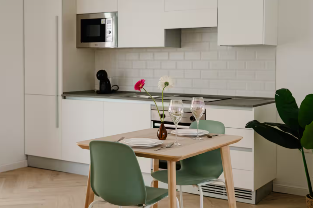 A modern kitchen in a stylish vacation rental features white cabinets, a microwave, and a coffee maker. A cozy dining table for two is set with plates, wine glasses, and flowers. Green chairs and a large plant complete this inviting short term rental space.