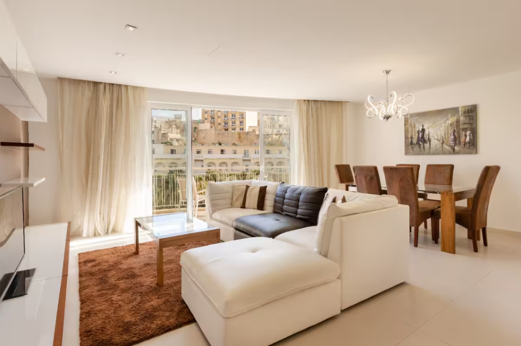 A modern living and dining area perfect for short term rentals, featuring a white sectional sofa, brown rug, dining table with six chairs, large windows with light curtains, city views, a chandelier, and stylish wall art.