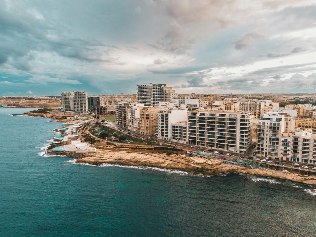 Aerial view of a coastal city with modern buildings and vacation rentals along the rocky shoreline, bordered by turquoise sea under a cloudy sky.