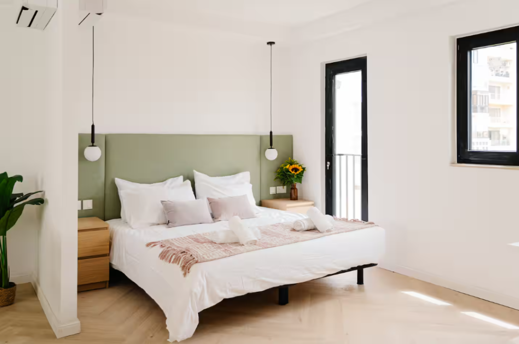 A modern, bright bedroom ideal for vacation rentals, featuring a green headboard, neatly made bed with white bedding, wooden nightstands, pendant lights, a potted plant, and sunflowers in a vase by black-framed windows.