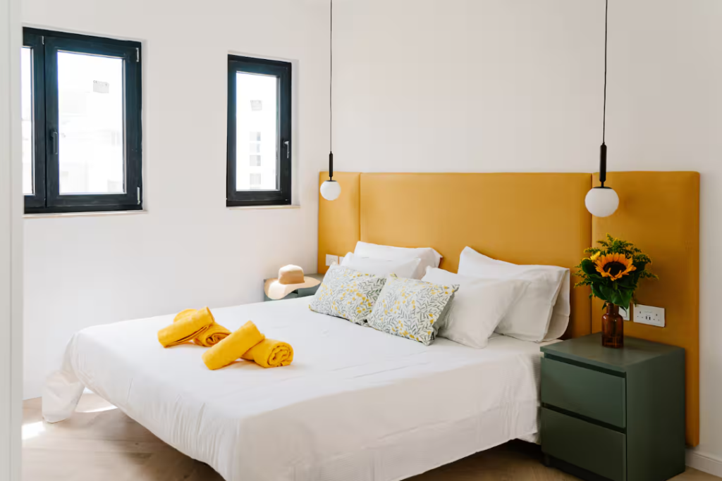 A bright, modern bedroom ideal for short term rentals, featuring a yellow headboard, white bedding, patterned pillows, and two yellow towels. Sunlight streams through two windows onto a green nightstand with sunflowers and stylish pendant lights above.