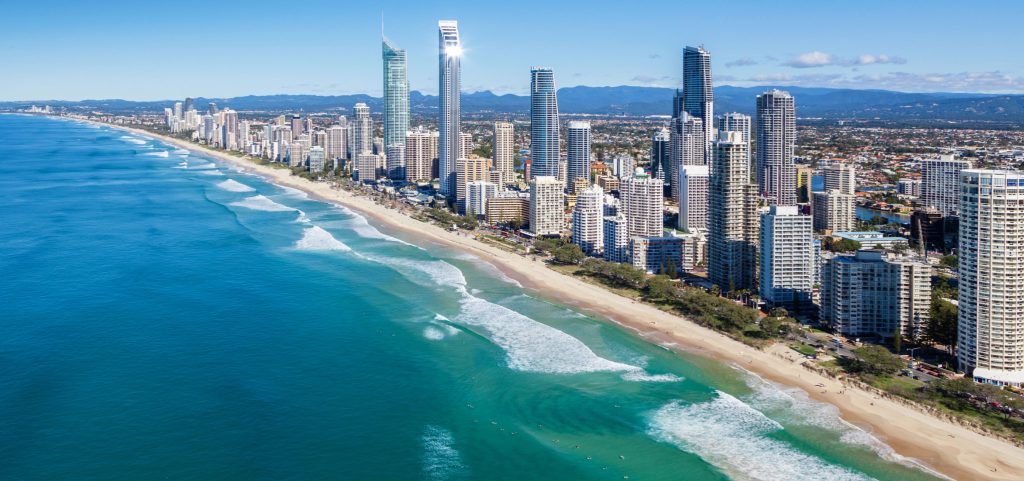 Aerial view of a city skyline with tall modern buildings, sandy beach, and turquoise waves—perfect for vacation rentals or short term rentals—set against a clear blue sky and distant mountains.
