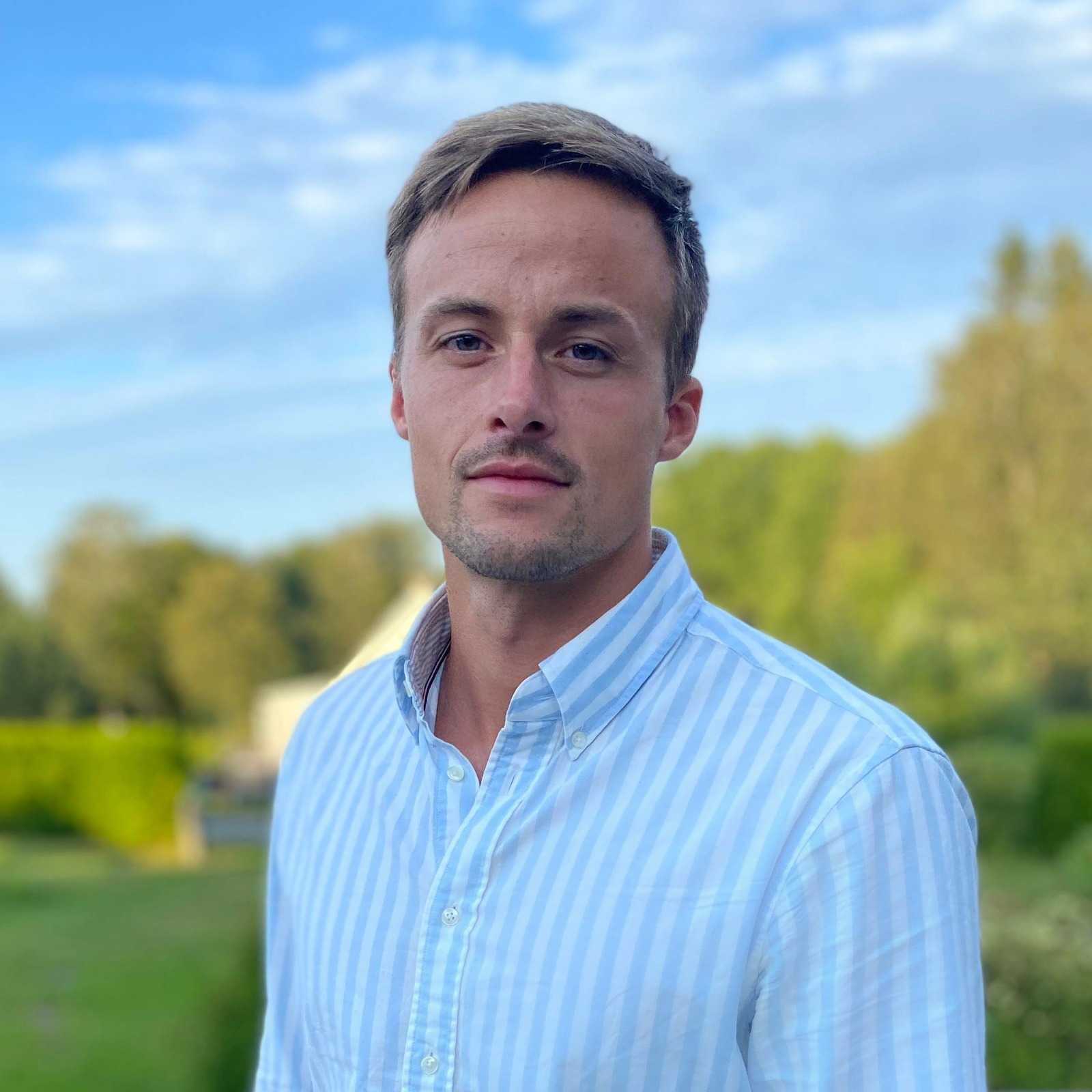 A man with short light brown hair and light facial hair stands outdoors in a light blue and white striped button-up shirt. Green trees and a blue sky suggest a peaceful setting, perfect for vacation rentals or exploring property management opportunities.