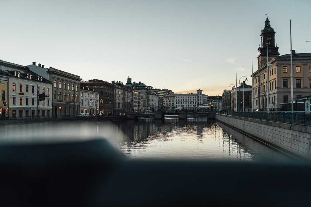 A calm river runs between historic European buildings at dusk, with reflections on the water and a clock tower visible on the right—an ideal setting for vacation rentals or short term rentals under a clear sky.