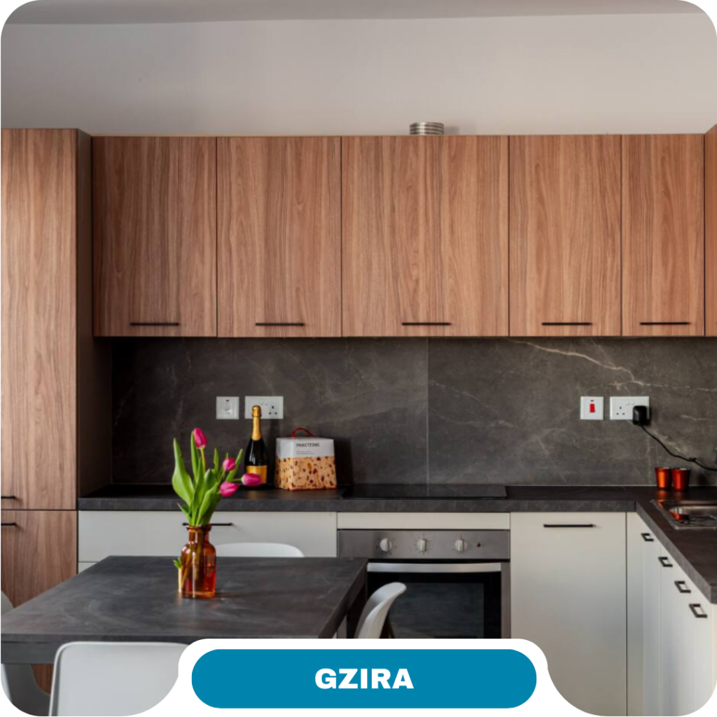 Modern kitchen with wood cabinets, grey countertops and backsplash, a built-in oven, white chairs, and a table with a vase of pink tulips—perfect for short term rentals. The location label at the bottom reads "GZIRA.