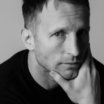 A man with short hair and a trimmed beard rests his chin on his hand, looking thoughtfully at the camera. The black and white image, with soft lighting, evokes the calm reflection needed in property management.