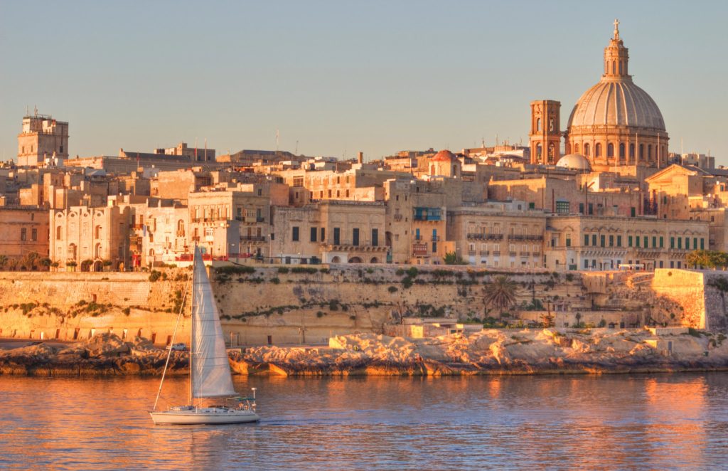 A sailboat glides on calm water at sunset in front of Valletta, Malta’s historic skyline, where golden limestone buildings and a domed cathedral create the perfect setting for memorable vacation rentals.