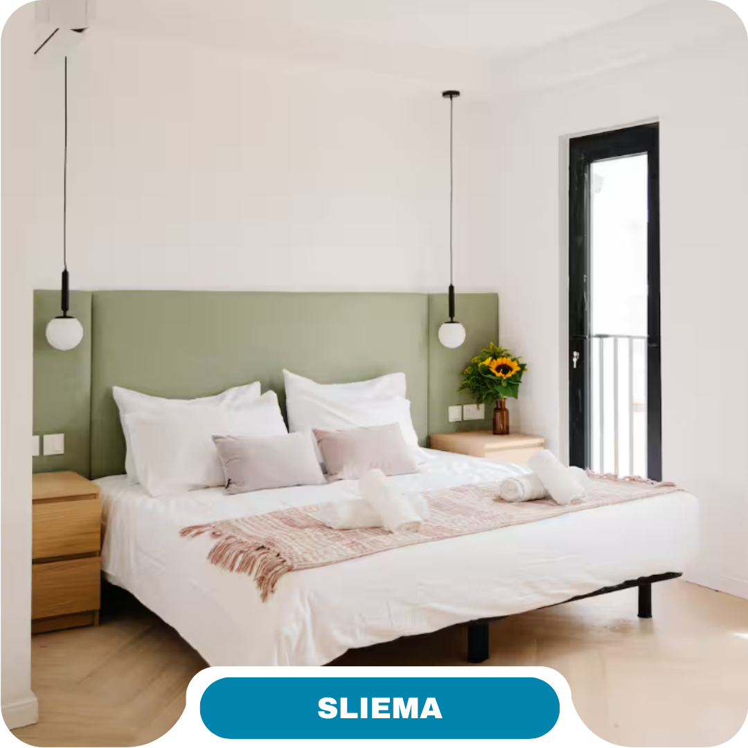 A bright, modern bedroom with a green headboard, double bed with white and pink bedding, stylish pendant lights—perfect for short term rentals or vacation rentals in Sliema. Wooden nightstands, sunflowers, and balcony access add charm.
