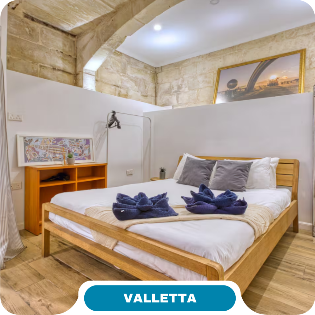 A cozy bedroom in Valletta with stone walls, a wooden bed, white bedding, gray and white pillows—perfect for short term rentals. Enjoy towel swans, a bedside table, wall art, and warm lighting for your next getaway.