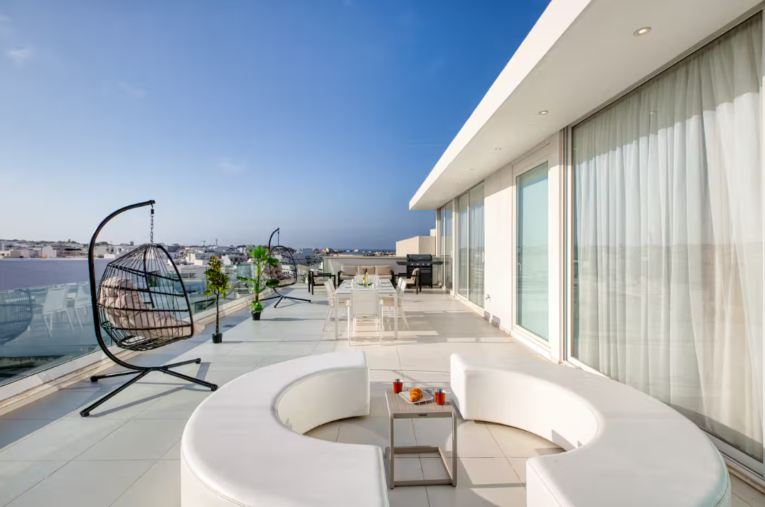 A spacious, modern rooftop terrace with white curved seating, hanging chairs, a dining table, and glass railings awaits—perfect for vacation rentals or short term rentals overlooking a cityscape under a clear blue sky.
