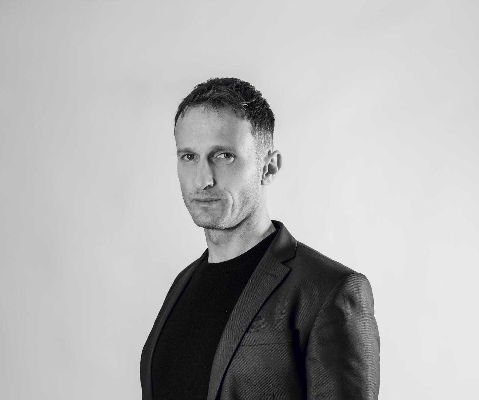 A man with short hair wearing a dark blazer and a black shirt stands against a plain, light background. He faces the camera with a neutral expression in this black-and-white photo.
