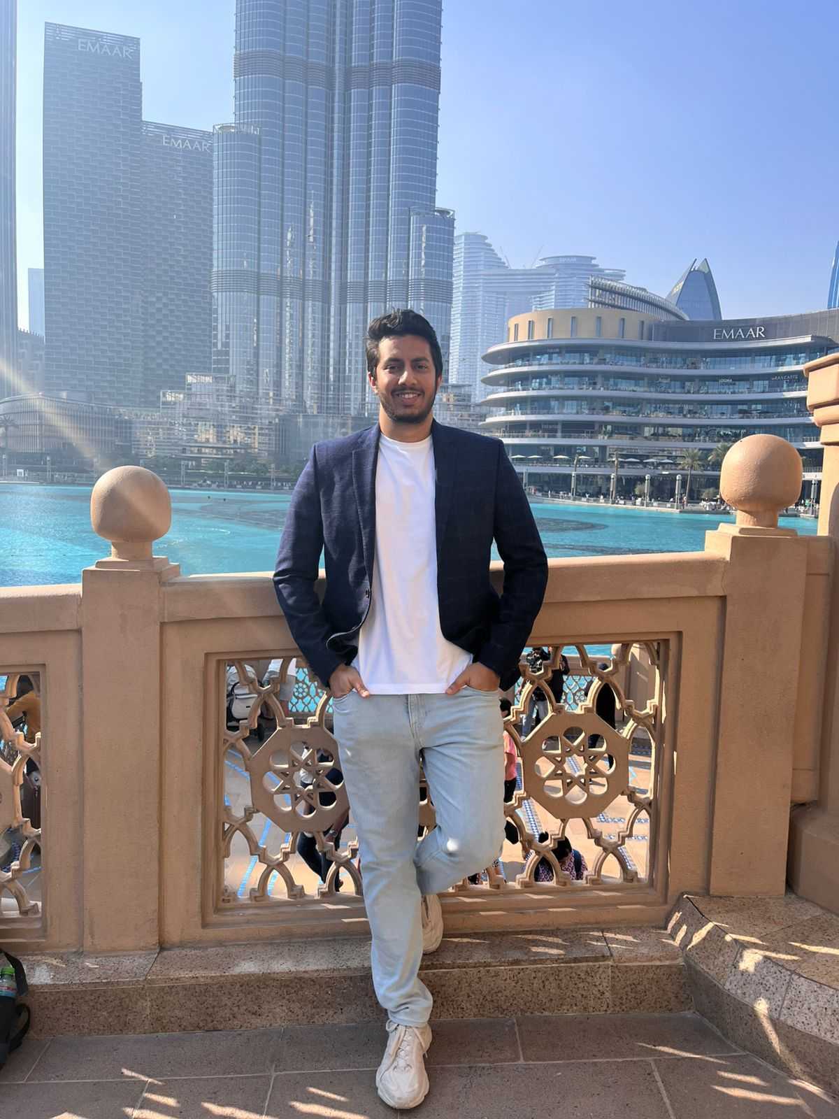 A man in a dark blazer, white shirt, and light jeans stands smiling by a decorative railing, with modern skyscrapers and the blue water of Dubai’s Burj Khalifa area visible in the sunny background.