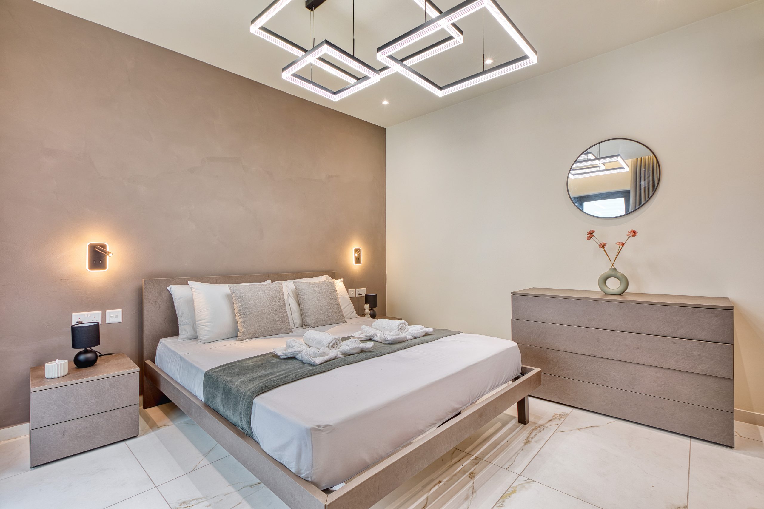 A modern bedroom in a luxury villa in Swieqi, Malta, featuring a large bed, neutral bedding, geometric ceiling lights, a round mirror above the dresser, and minimalist decor in soft beige and gray tones.