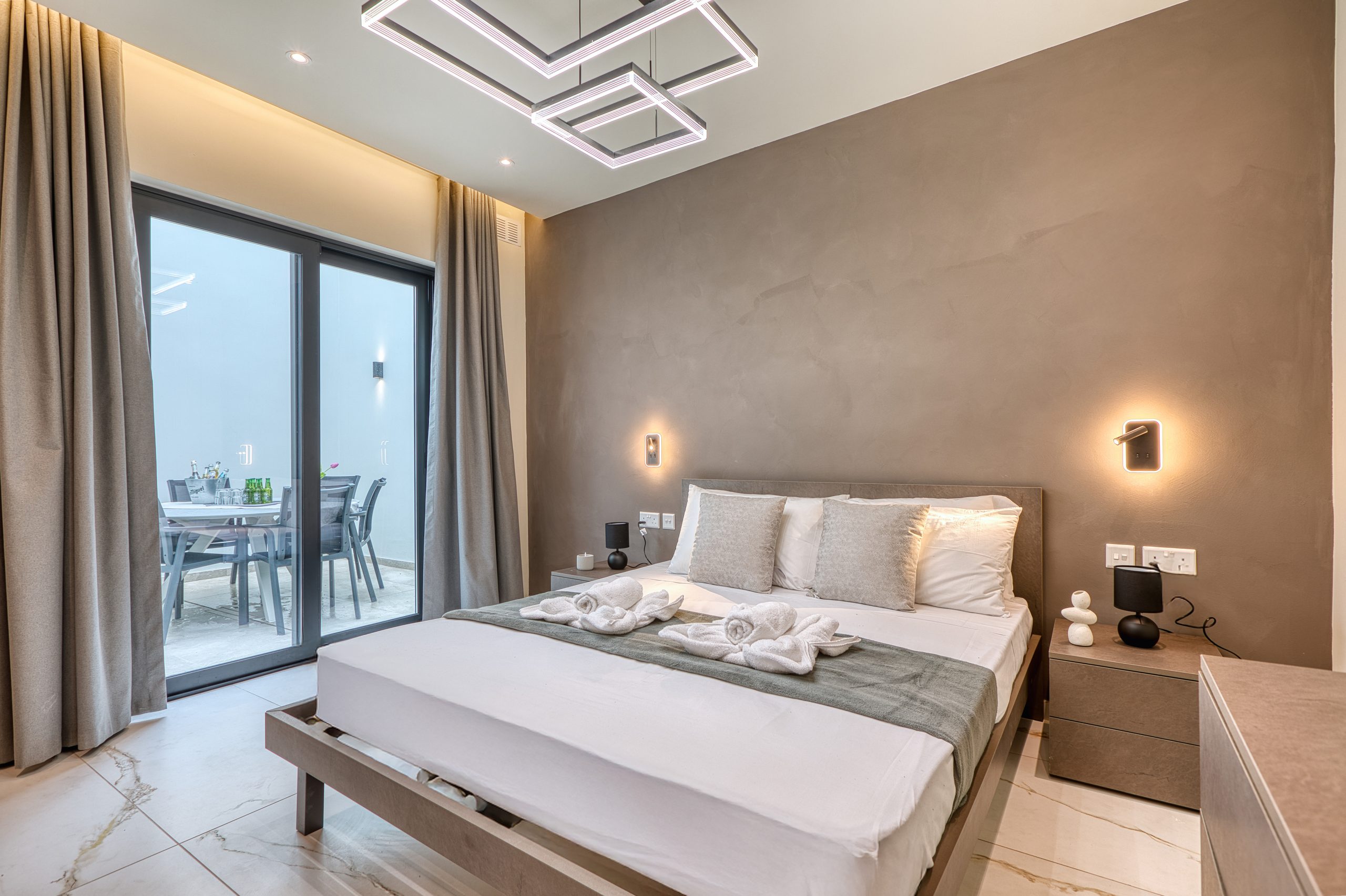 Modern bedroom in a luxury villa in Swieqi, Malta, with a large bed, minimalist decor, bedside tables, and wall-mounted lights. Sliding glass doors open to an outdoor patio. Neutral colors and soft lighting create a cozy feel.