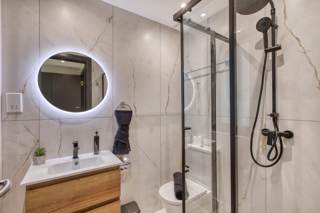 Modern bathroom in a luxury villa in Swieqi, Malta, featuring marble-patterned walls, a round backlit mirror above a wooden vanity with a small sink, glass shower enclosure with black fixtures, and a potted plant on the countertop.