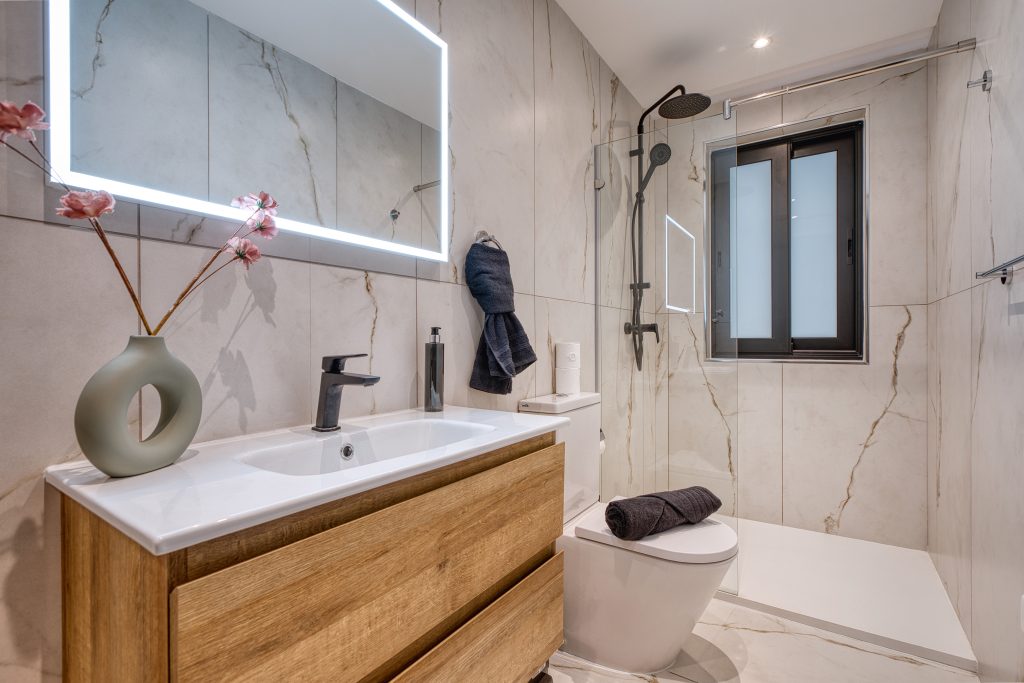 Modern bathroom in a Swieqi luxury villa featuring a wood vanity, vessel sink, large backlit mirror, glass shower with black fixtures, wall-mounted toilet, and window. Marble-style walls and floor complement pink flowers on the counter.