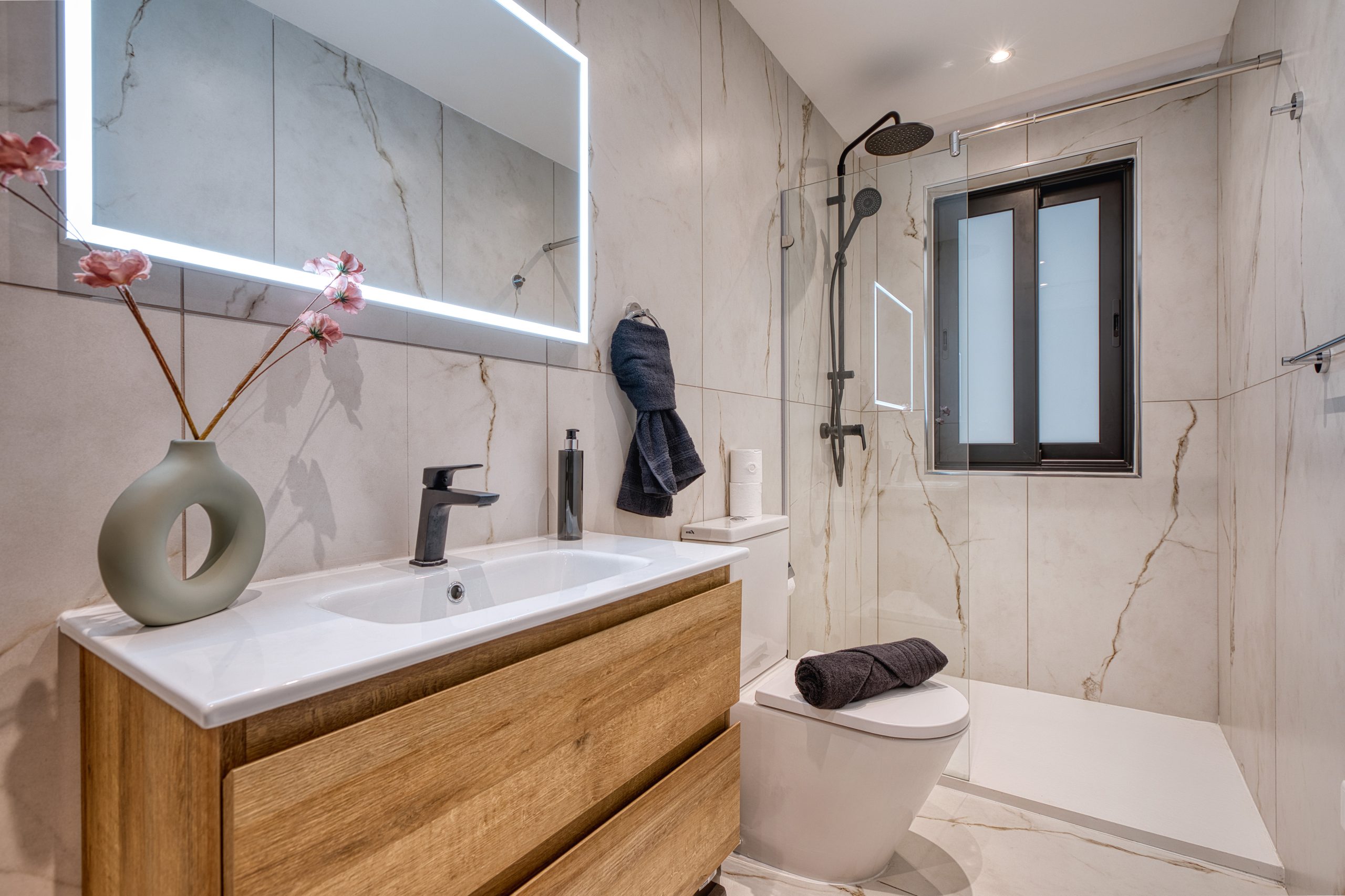 Modern bathroom in a Swieqi luxury villa featuring a wood vanity, vessel sink, large backlit mirror, glass shower with black fixtures, wall-mounted toilet, and window. Marble-style walls and floor complement pink flowers on the counter.