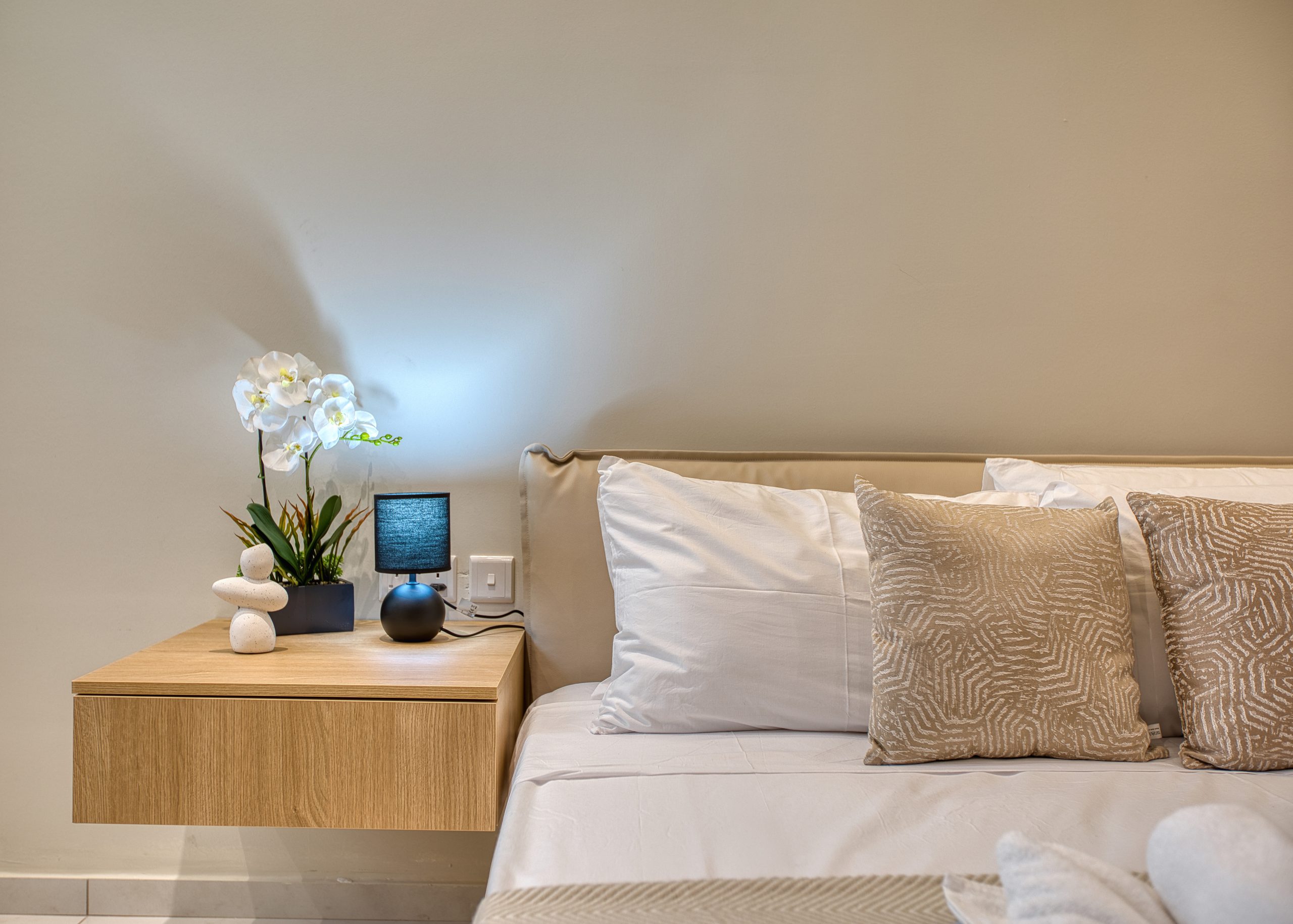 A modern bedroom in a luxury villa in Swieqi, Malta, featuring a floating wooden nightstand, white orchid plant, small black lamp, and stacked stones beside a neatly made bed with white and beige patterned pillows.
