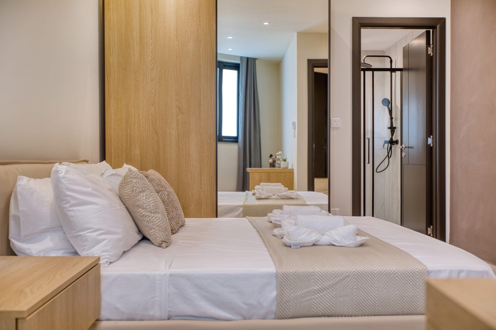 A modern, neatly made bed with white and beige linens awaits in this Swieqi luxury villa. Wooden furniture, a large mirror, and an open door to a bathroom with shower complete the scene, as natural light pours in through the window.