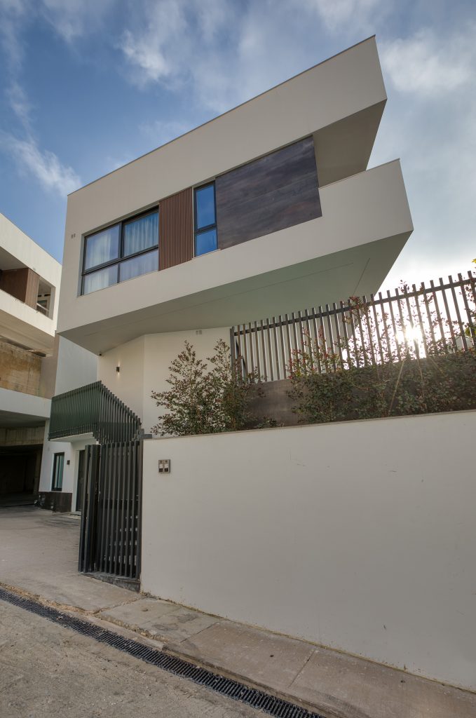 A modern two-story luxury villa in Swieqi, Malta, featuring a geometric design, white exterior walls, large windows, and a wood accent panel. A metal fence and plants line the front of the property under a partly cloudy sky.