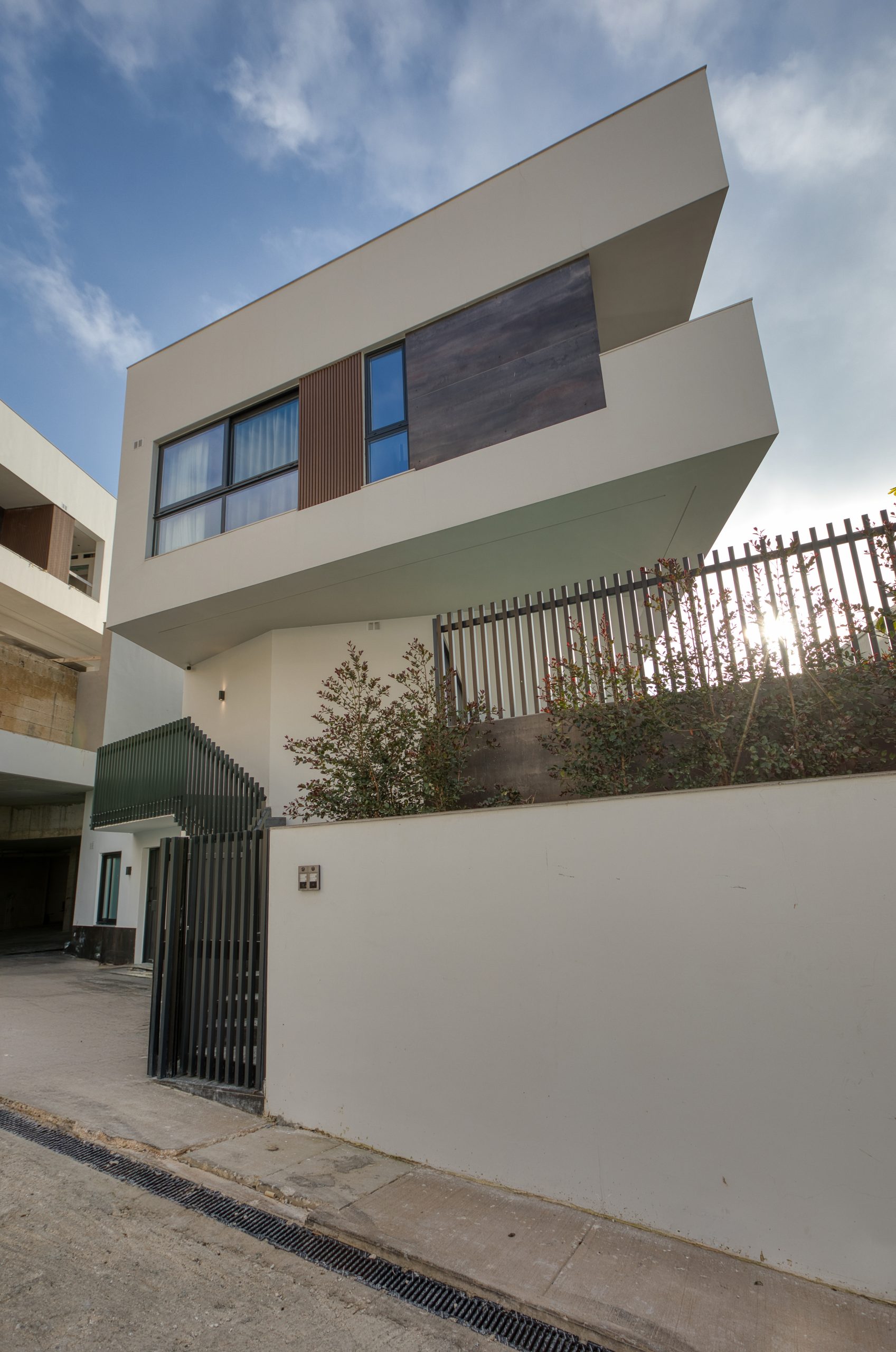 A modern two-story luxury villa in Swieqi, Malta, featuring a geometric design, white exterior walls, large windows, and a wood accent panel. A metal fence and plants line the front of the property under a partly cloudy sky.