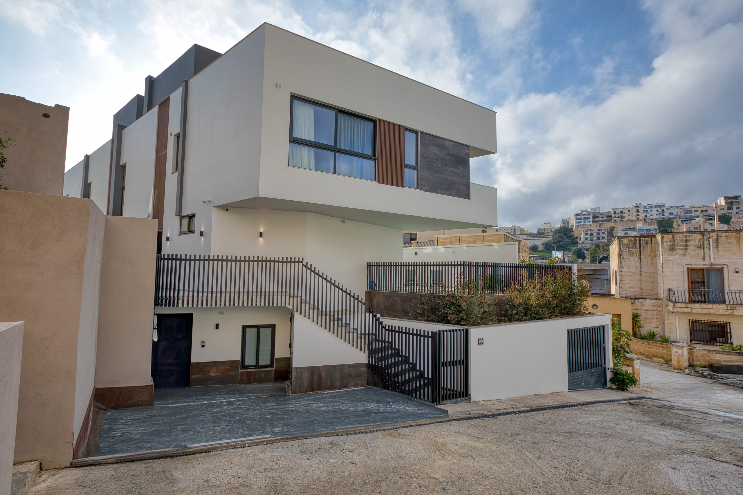 Modern two-story white Luxury Villa with large windows, an exterior staircase, and a gated driveway, set on a sloped street in Swieqi, Malta, surrounded by other buildings and greenery under a partly cloudy sky.