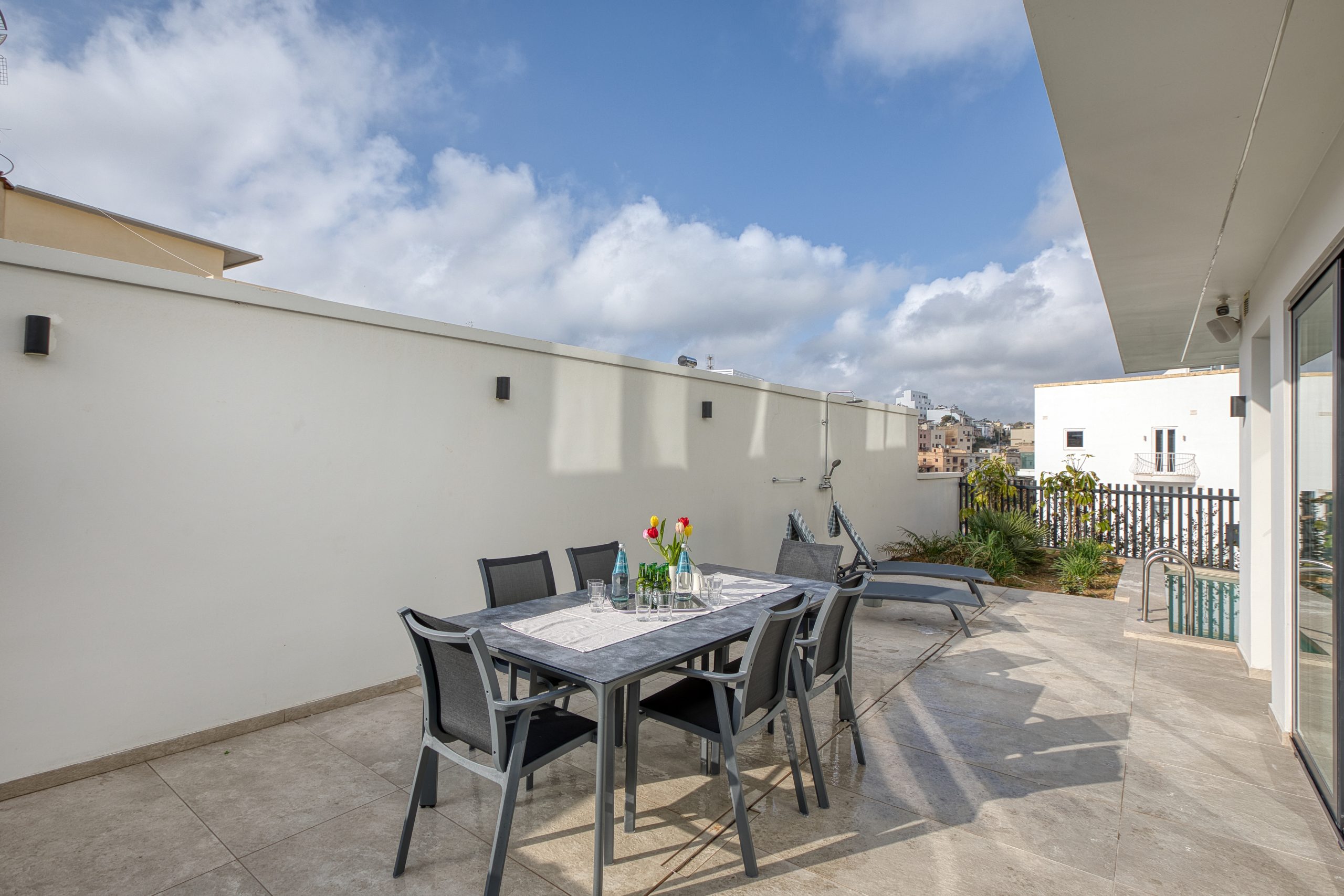 A modern outdoor patio at a luxury villa in Swieqi, Malta, with a table set for four, black chairs, a vase of tulips, and lounge chairs against a white wall. The area is sunny with blue sky and scattered clouds.