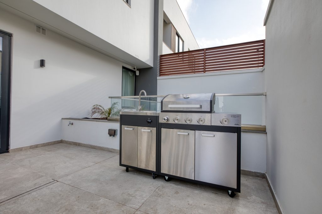 A modern stainless steel outdoor grill with a sink stands on a spacious, clean patio surrounded by white walls and a wooden privacy screen in this luxury villa in Swieqi, Malta.