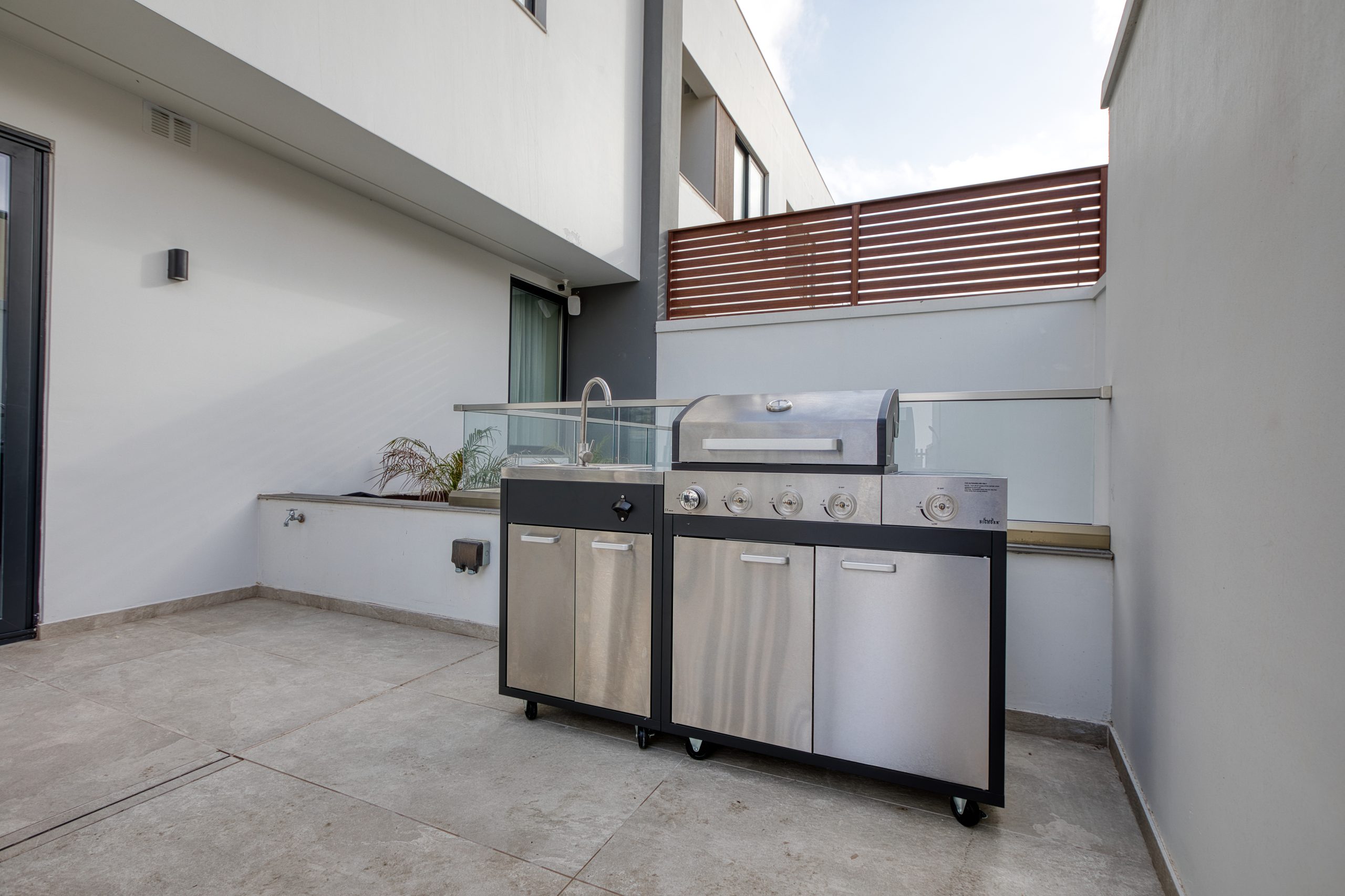 A modern stainless steel outdoor grill with a sink stands on a spacious, clean patio surrounded by white walls and a wooden privacy screen in this luxury villa in Swieqi, Malta.
