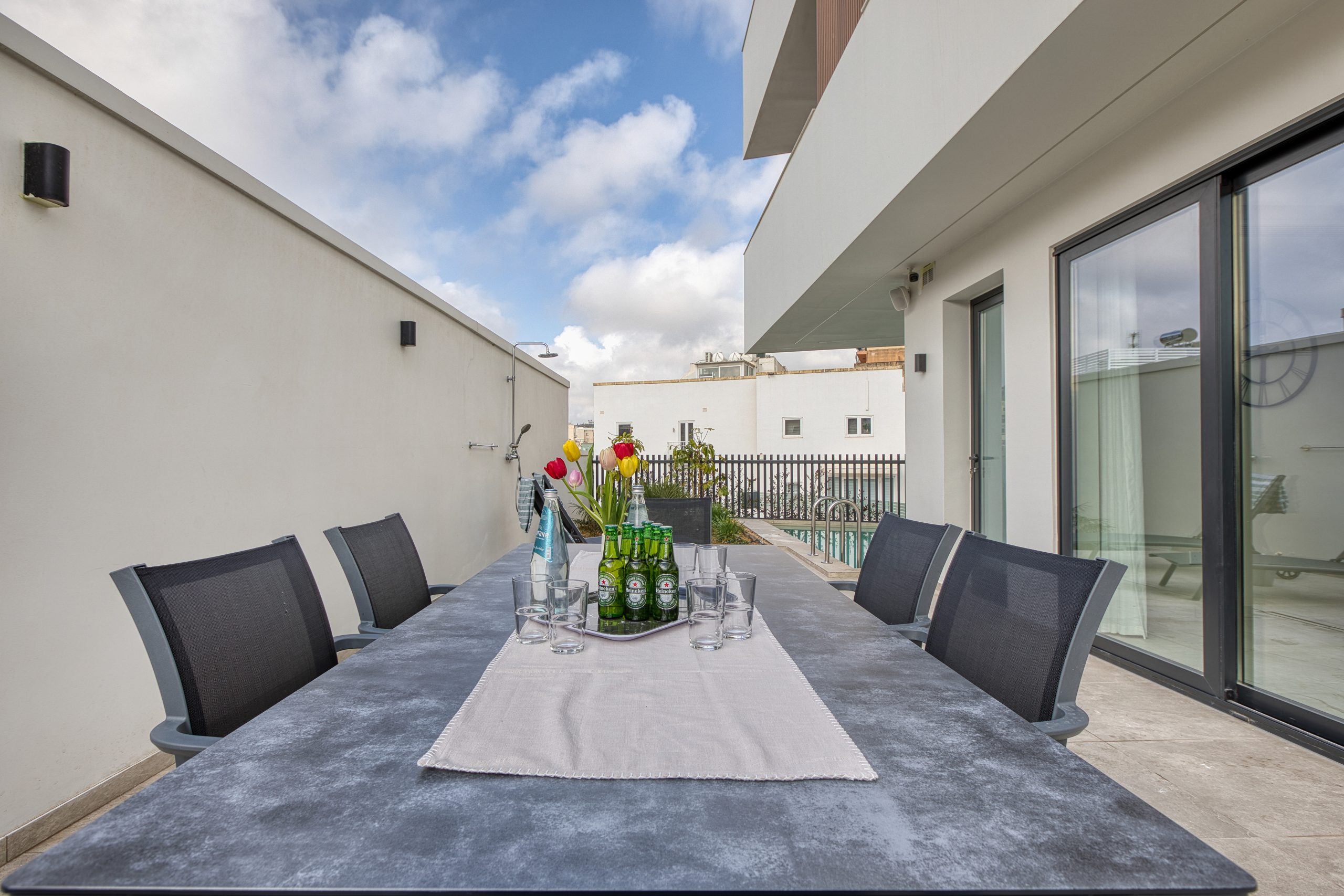 A modern outdoor patio at a luxury villa in Swieqi, featuring a long dining table set with drinks, glasses, and vibrant flowers, surrounded by black chairs—all framed by white walls and the blue Malta sky punctuated with clouds.