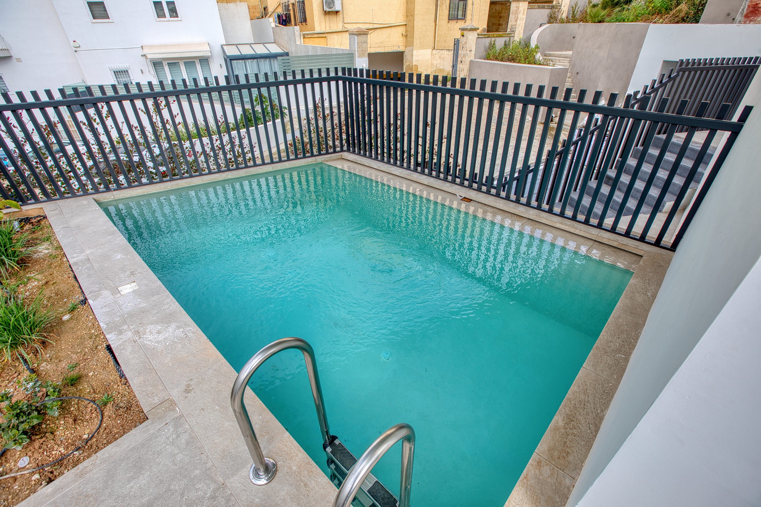 A small rectangular backyard swimming pool with clear blue water, surrounded by beige tiles and a black metal fence, sits beside a building and lush plants in a luxury villa in Swieqi, Malta. A metal ladder leads invitingly into the pool.
