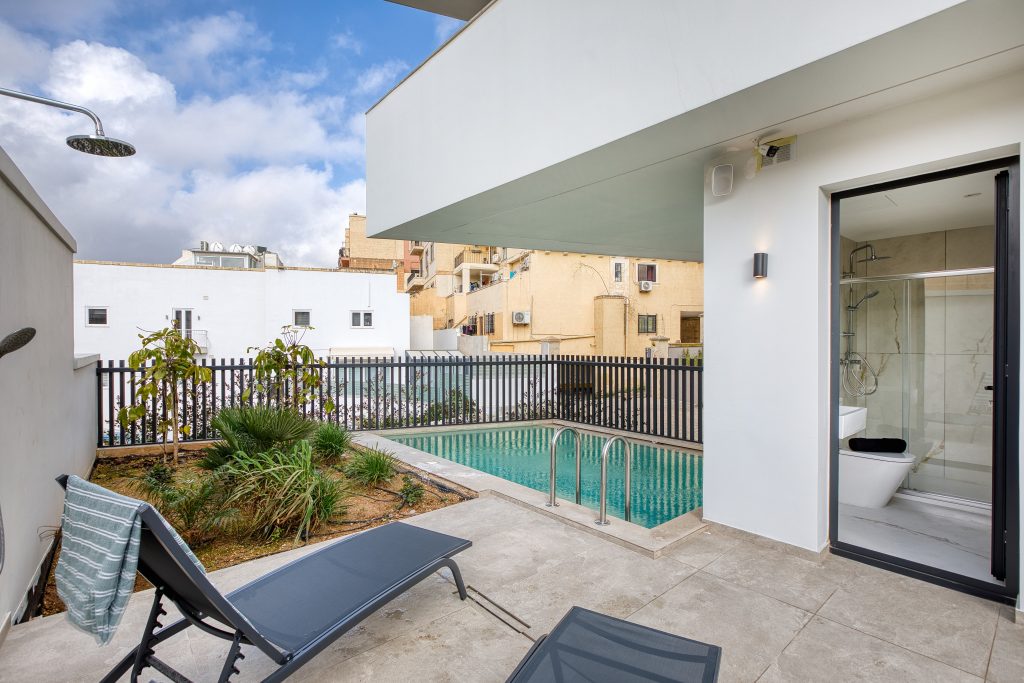 A modern luxury villa patio in Swieqi, Malta, features two lounge chairs, a towel, a small garden, an outdoor shower, and a rectangular swimming pool beside a white building with glass doors. Urban buildings are visible in the background.