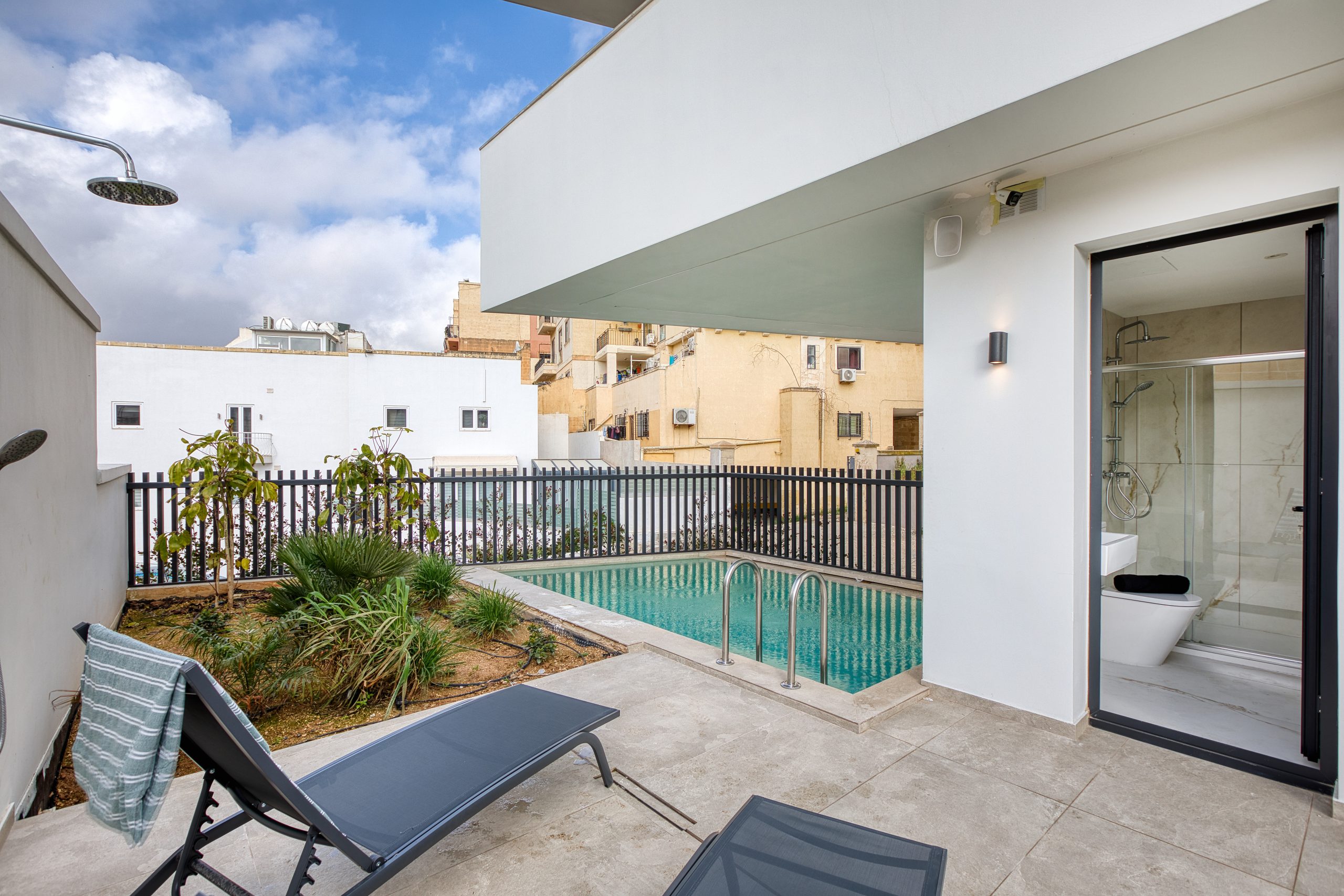 A modern luxury villa patio in Swieqi, Malta, features two lounge chairs, a towel, a small garden, an outdoor shower, and a rectangular swimming pool beside a white building with glass doors. Urban buildings are visible in the background.