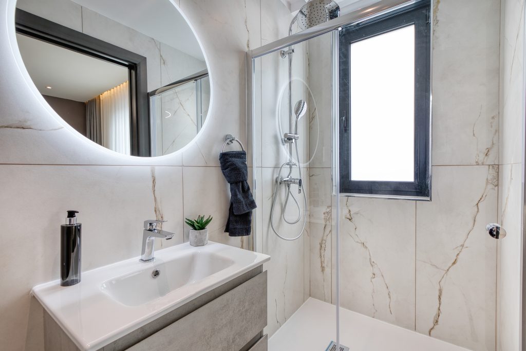 Modern bathroom in a luxury villa in Swieqi, Malta, featuring marble walls, a glass shower enclosure, a rectangular sink with mirror above, black soap dispenser, small potted plant, and a window providing natural light.