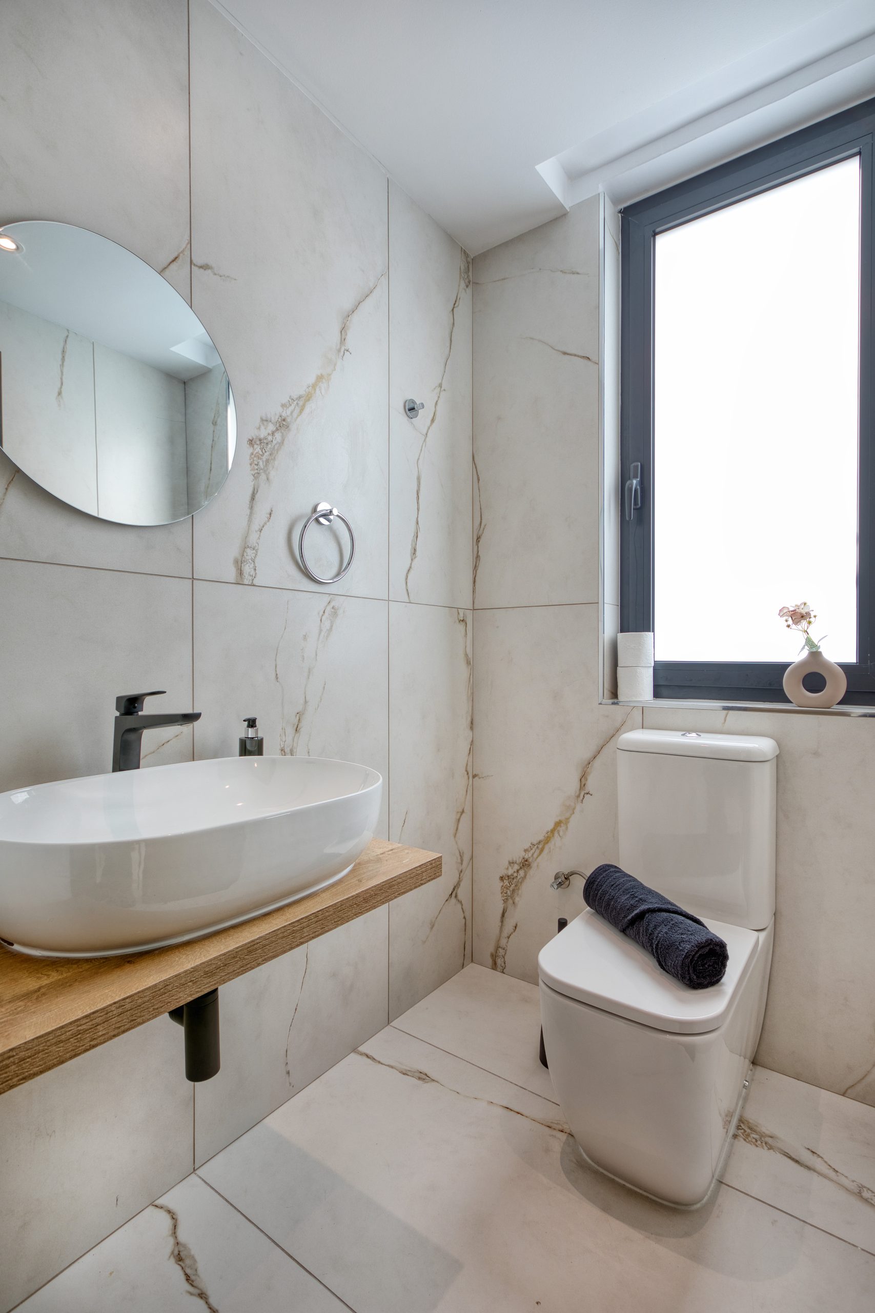 Modern bathroom in a luxury villa in Swieqi, Malta, featuring marble-patterned tiles, a round mirror, vessel sink on a wooden counter, toilet with a rolled dark towel, and a large frosted window with a small decorative vase.