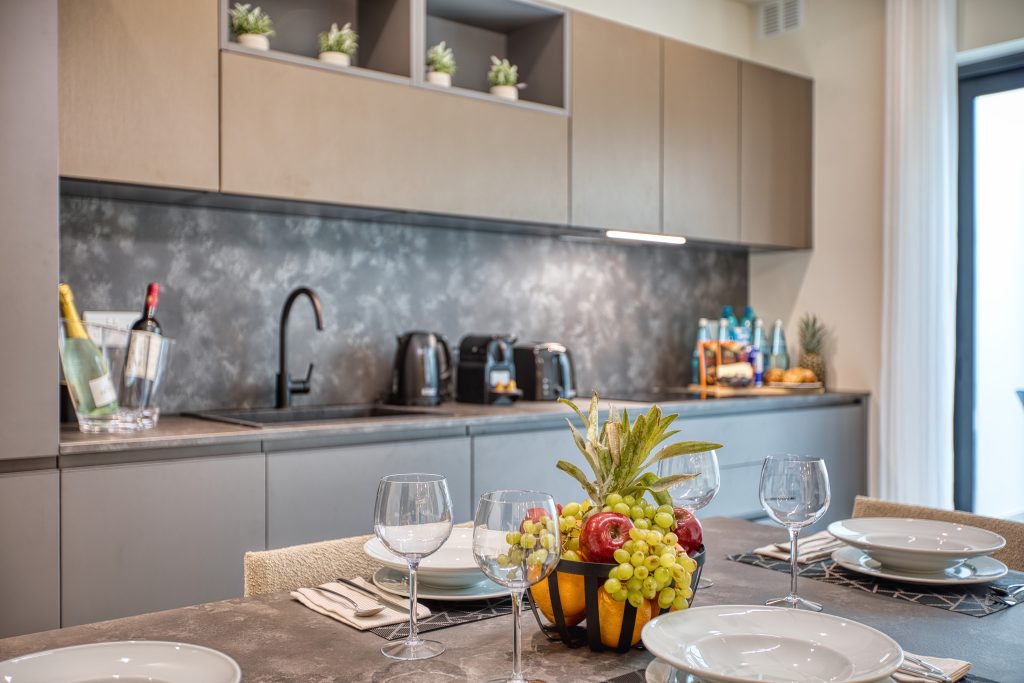 Modern kitchen with gray cabinets, black sink, and appliances in a luxury villa in Swieqi, Malta. The countertop displays wine, bottles, and a fruit basket. A dining table is set with plates, glasses, and a centerpiece of fresh fruit.
