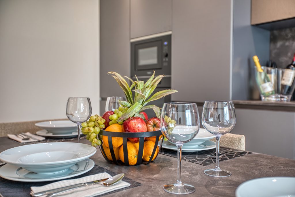A modern dining table set with white plates, bowls, and wine glasses graces this luxury villa in Swieqi, Malta. A black fruit basket overflowing with apples, oranges, grapes, and a pineapple centerpiece sits amid a kitchen with wine bottles in the background.
