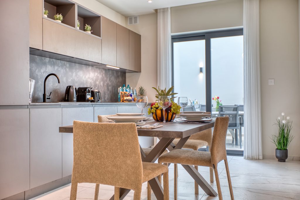 Modern kitchen and dining area in a luxury villa with beige cabinets, sleek countertop, and appliances. A table set for four with tan chairs and a plant centerpiece stands near large glass doors in Swieqi, Malta, leading to an outdoor patio.